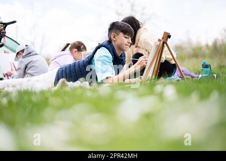 Glückliche junge Familie, Mutter und Kinder, die Spaß haben und sich im Freien auf Picknick-Decken-Malerei im Garden Spring Park amüsieren, Entspannung. Stockfoto