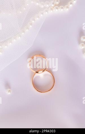 Zwei goldene Eheringe auf schickem weißem Satinhintergrund mit Perlenkette. Vertikaler Hochzeitshintergrund. Layout für Design Stockfoto