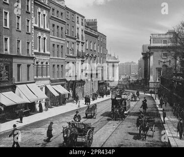 Ein Foto aus dem späten 19. Jahrhundert von Grafton Street, Dublin City Centre, Irland am College Green End, ursprünglich eine modische Wohnstraße mit einigen kommerziellen Aktivitäten. Bis 1849 wurde es immer öfter veraltet, dann wurden Ende des 19. Jahrhunderts eine Reihe von Einzelhandelsgeschäften gebaut und mehrere langjährige Unternehmen etablierten sich auf der Straße, wie zum Beispiel die Kaufhäuser Switzer's und Brown Thomas. Die Juwelierweiber wurden 1869 eröffnet. Während des 20. Jahrhunderts wurde es bekannt für das Kaffeehaus Bewley's, für die Geschäfte im mittleren und gehobenen Marktsegment und als beliebter Ort für Stockfoto