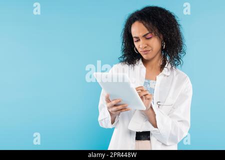 Verärgerte afroamerikanische Frau in weißem Hemd, die ein digitales Tablet benutzt, isoliert auf blauem Stock-Bild Stockfoto