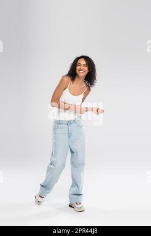 Volle Länge einer fröhlichen afroamerikanischen Frau in blauen Jeans und Turnschuhen, die auf grauem Hintergrund tanzen, Stockbild Stockfoto