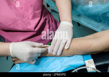 Platzierung eines Kathetertropfers im Arm eines auf dem Operationstisch liegenden Patienten. Führen Sie die Nadel in sterile Handschuhe mit den Händen des Arztes in den Arm des Patienten ein. Stockfoto