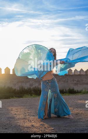 Orientalische Tänzerin in Blau mit Isis-Flügeln der gleichen Farbe, die bei Sonnenuntergang auf einem arabischen Denkmal tanzt Stockfoto