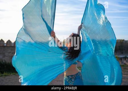 Orientalische Tänzerin in Blau mit Isis-Flügeln der gleichen Farbe, die bei Sonnenuntergang auf einem arabischen Denkmal tanzt Stockfoto