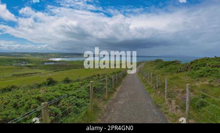 Wander- und Wanderwege in den grünen Hügeln des Giant's Causeway, Irland, mit Blick auf das Meer, blauem Himmel mit Wolken und einem irischen Dorf im Hintergrund Stockfoto