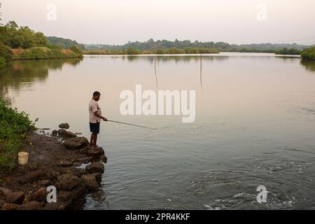 Goa, Indien - 19. Dezember 2022 : ein einsamer Fischer, der am Ufer eines Flusses steht und spät am Abend mit einer Angelrute Fische fängt. Stockfoto