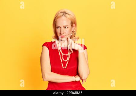 Porträt einer blonden Frau in rotem Kleid, die in die Kamera schaut, mit nachdenklichem Gesichtsausdruck auf gelbem Hintergrund. Ich mache einen Geschäftsplan Stockfoto