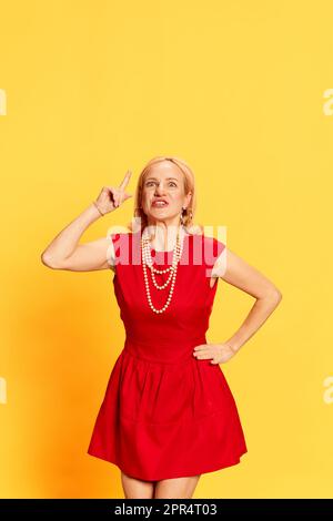 Eine attraktive blonde Frau trägt ein schickes rotes Kleid und kreiert gerade gute Ideen mit gelbem Studiohintergrund. Einblicke Stockfoto