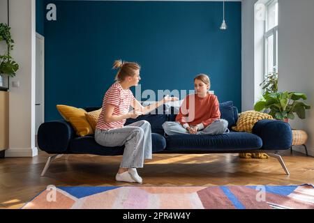 Aufgebrachtes Teenager-Mädchen, das auf dem Sofa sitzt und zuhört, wie die wütende Mutter sie wegen schlechter Schulnoten schimpft Stockfoto