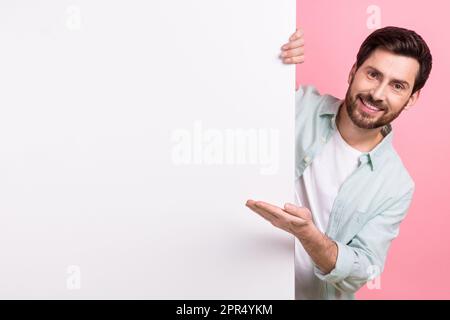 Foto eines jungen, lustigen Geschäftsmannes, der sich hinter einer großen Plakette versteckt, zeigt die Hand mit dem neuen Produktnamen isoliert auf pinkfarbenem Hintergrund Stockfoto