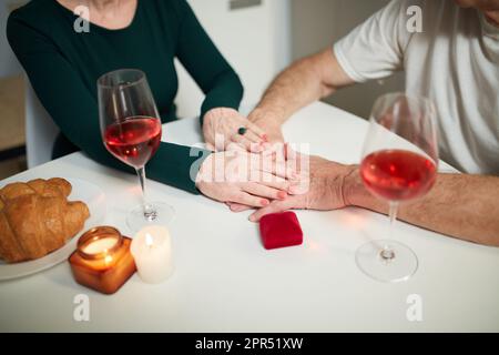 Nahaufnahme von Pärchenhänden, die Unterstützung und Liebe geben. Seniorenpaar mit romantischem Abendessen zu Hause in der Küche mit Wein und Geschenken Stockfoto