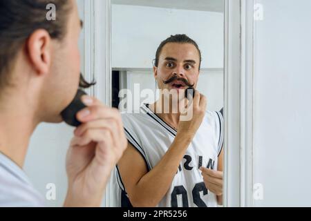 Junge Latina-geschlechtliche, nicht binäre Person, die in ihrem Zimmer steht und in den Spiegel schaut, während sie Make-up mit einem Schwamm aufträgt, Schönheitskonzept, konzentriere dich auf das Ba Stockfoto