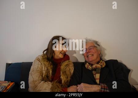 Lächelnd, glücklich, lachend, pensioniertes Paar, das auf dem Sofa sitzt, die Fäuste schlägt - und Winterkleidung drinnen trägt Stockfoto