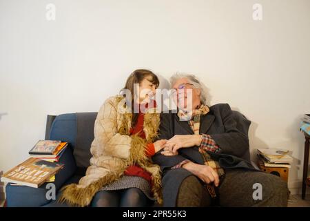 Lächelndes, glückliches, lachendes und positives pensioniertes Paar, das auf dem Sofa sitzt, Hände hält und sich in die Augen schaut - in Winterkleidung drinnen Stockfoto