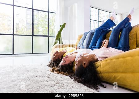 Drei junge, multiethnische Frauen, die Spaß haben, während sie zusammen auf dem Sofa lachen Stockfoto