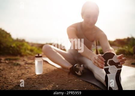 Lächelnde junge weiße Dame in Sportkleidung, die sich mit einer Flasche Wasser in der Natur am Strand die Beine streckt Stockfoto