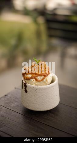 Kleine oder eine Schaufel Vanilleeis in einer Tasse, mit braunem Zucker und Karamellsauce, Seitenansicht auf Holztisch Hintergrund Stockfoto