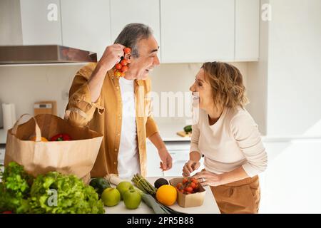 Fröhliche ältere Ehepartner, die nach der Ankunft aus dem Supermarkt Gemüse aus der Papiertüte nahmen und Spaß in der Küche hatten Stockfoto