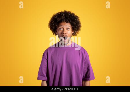 Positiv aufgeregter, schockierter erwachsener, afroamerikanischer Lockenkopf in lila T-Shirt mit offenem Mund Stockfoto