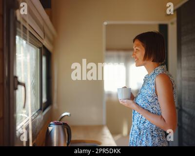 Junge Frau genießt morgens eine Tasse Kaffee in ihrer Küche. Minimalismus. Wir ziehen ein. Allein zu leben. Charmantes Wohnwagenhaus Stockfoto