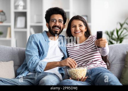 Lächelnde gemischtrassige Ehepartner, die zu Hause auf der Couch sitzen und fernsehen Stockfoto