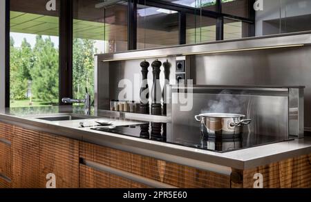 Stahltopf mit kochenden und dampfenden Speisen in moderner Küche aus Holz und Edelstahl. Kochen. Stockfoto