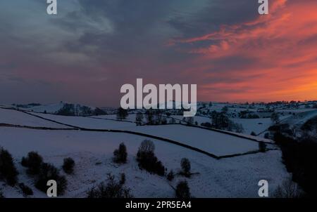 Die schneebedeckte Landschaft von Staffordshire im Peak District, kurz nach Sonnenuntergang mit noch etwas Rot am Himmel. Ackerland, Bäume und Trockenmauern Stockfoto