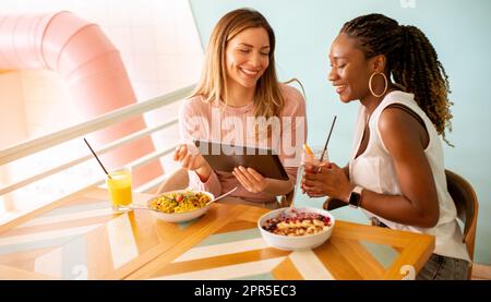 Zwei hübsche junge schwarze und weiße Frau, die sich amüsiert, frische Säfte trinkt und ein gesundes Frühstück im Café hat Stockfoto