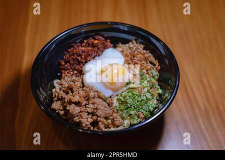 Eine Schüssel Chili Pan Mee mit halbgekochtem Ei, Sardellenfisch, getrocknetem Chili und Hühnerfleisch auf einem Holztisch Stockfoto