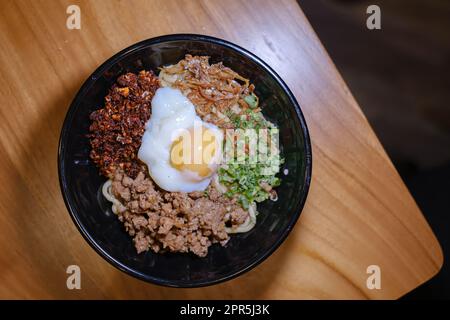 Flach liegend oder von oben, Aufnahme einer Schüssel Chili Pan Mee mit halbgekochtem Ei, Sardellenfisch, getrocknetem Chili und Hühnerfleisch auf einem Holztisch Stockfoto