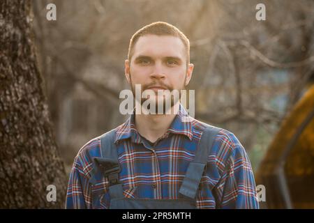 Ein Bauer von europäischem Aussehen, ein ländliches Porträt im Sonnenlicht, mit Bart, Hemd und Overall, blickt auf die Kamera draußen. Stockfoto