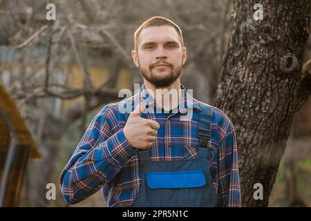 Bauer befriedigte das landporträt mit dem bart, Hemd und Overall, die auf die Kamera blickten und im Freien eine coole Geste mit dem Handfinger zeigten. Stockfoto