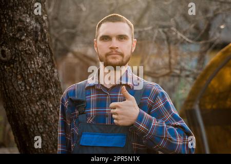 Der Bauer erfüllte das europäische Erscheinungsbild des ländlichen Portraits bei Sonnenlicht mit Bart, Hemd und Overall, die in die Kamera blickten und eine coole Geste mit Handfink zeigten Stockfoto