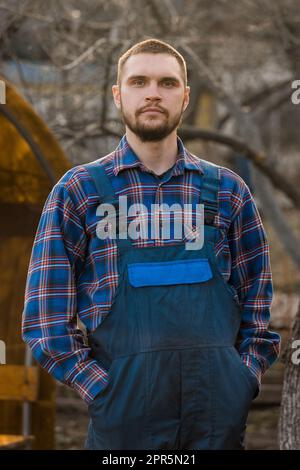 Bauer ernstes europäisches Aussehen männliches ländliches Porträt mit Bart, Hemd und Overall mit Händen in Taschen, die draußen auf die Kamera schauen. Stockfoto