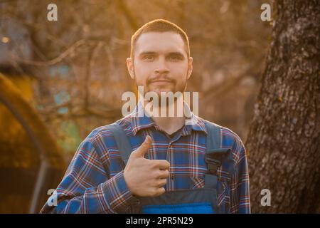 Bauer befriedigte das landporträt des europäischen Erscheinungsbildes bei Sonnenuntergang mit Bart, Hemd und Overall, die in die Kamera blickten und mit dem Finger eine coole Geste zeigten Stockfoto