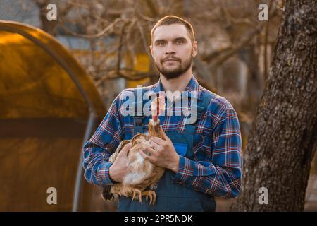 Bauer hübsches europäisches Aussehen männliches ländliches Porträt bei Sonnenuntergang mit Bart, Hemd und Overall mit weißem Huhn in den Armen draußen CO Stockfoto