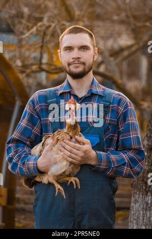 Bauer hübsches europäisches Aussehen männliches ländliches Porträt mit Bart, Hemd und Overall mit weißem Huhn in den Armen draußen auf dem Land. Stockfoto