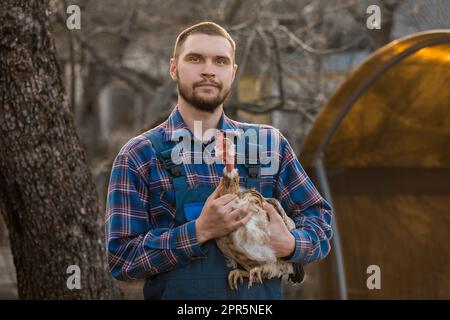 Bauer hübsches europäisches Aussehen männliches ländliches Porträt auf dem Land mit Bart, Hemd und Overall mit weißem Huhn in den Armen draußen Stockfoto