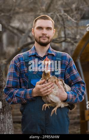 Bauer hübsches europäisches Aussehen Weißer Mann Landporträt mit Bart, Hemd und Overall mit weißem Huhn in den Armen draußen CO Stockfoto