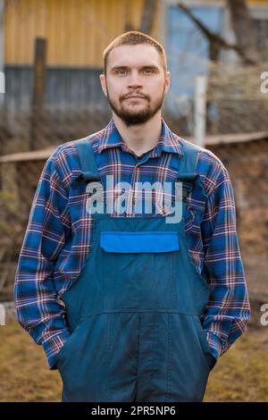 Bauer hübsches europäisches Aussehen männliches ländliches Porträt mit Bart, Hemd und Overall mit Händen in Taschen, Blick auf die Kamera im Freien auf dem Land. Stockfoto