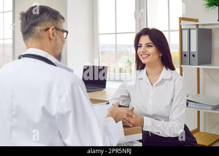 Fröhliche junge brünette Frau bei einem Arzttermin mit Brille, die Einträge in die Patientenakte macht. Stockfoto