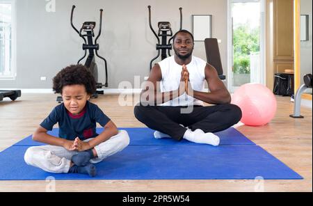 Junger, kurzer, lockiger, schwarzer Vater mit Schnurrbart und Bart meditiert auf Yogamatte vor dem Training. Der Junge sitzt unglücklich vorne. Morgengymnastik, Achtsamkeitskonzept. Stockfoto