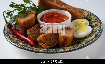 Jachnun oder Jahnun, jüdisches Gebäck aus dem jemenitischen Raum, serviert mit frischer geriebener Tomate und gekochtem Ei und Zhug, die von den Adeni-Juden stammen, und traditionell serviert am Sabbat-Morgen in Israel. Stockfoto