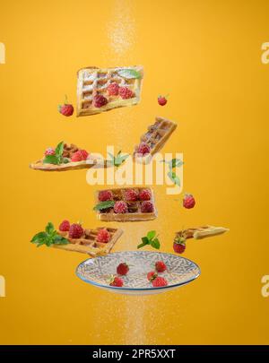 Gebackene belgische Waffeln, Himbeeren und Minzblätter schweben auf gelbem Hintergrund. Darüber Puderzucker streuen Stockfoto