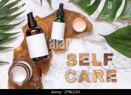 Self Care-Text neben Kosmetikflaschen und Monstera Leaves Draufsicht, Modell Stockfoto
