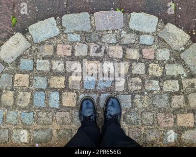 Zwei schwarze Schuhe und Beine auf einer Kopfsteinpflaster-Oberfläche - Draufsicht Konzept. Stockfoto