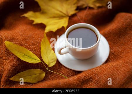 Stillleben mit schwarzem Kaffee in einer weißen Tasse, die auf einem warmen, gemütlichen Pullover steht, Draufsicht, Dekoration mit gelben Blättern im Hintergrund. Stockfoto