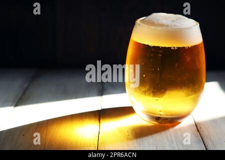 Ein schäumendes Bier, das in ein kurzes, abgerundetes Glas gegossen wurde, erstrahlt vom starken Nachmittagslicht Stockfoto