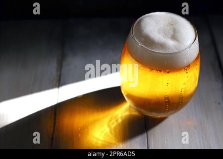 Ein schäumendes Bier, das in ein kurzes, abgerundetes Glas gegossen wurde, erstrahlt vom starken Nachmittagslicht Stockfoto