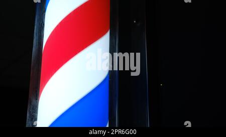 Ein altmodisches Barbier-Shop-Schild, Symbol. Runde Polbeleuchtung im Dunkeln. Altmodisches rotierendes LED-Pollicht mit roten weißen blauen Streifen in der Nähe des Männer-Friseursalons. Werbung für ein kleines Unternehmen im Freien. Friseur. Stockfoto